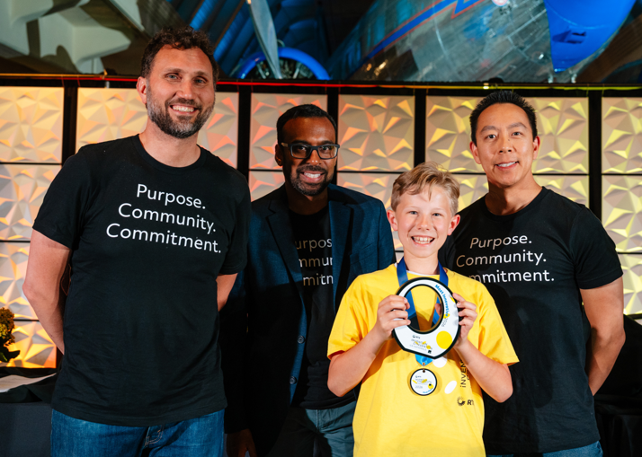 A boy in a yellow shirt poses with a white trophy award in front of three men
